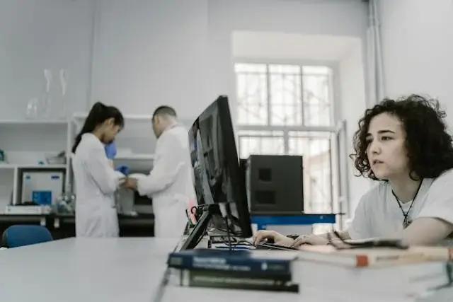 Synapse Lab Billing Services Three healthcare professionals in a laboratory wearing white coat, one is facing the computer with books on the table and the other two is working together.