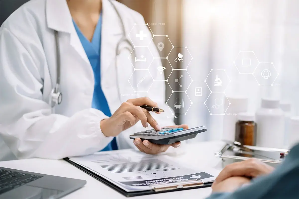A healthcare professional sitting on her table wearing blue scrub, white coat and a stethoscope on her shoulder, holding a pen and a calculator with a laptop and clipboard on her table